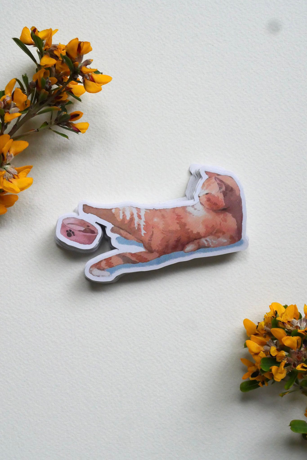 Watercolour sticker of a ginger cat lying down and stretching one paw towards a pink food bowl on a cream background with sprigs of yellow flowers