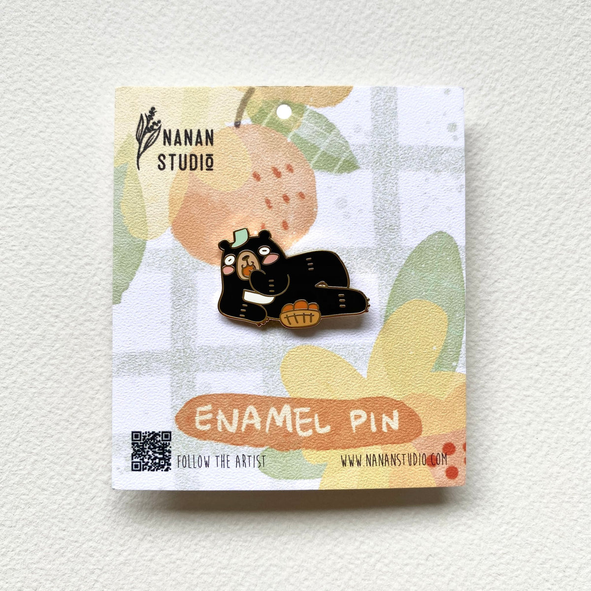 Principal Bear enamel pin, a Formosan black bear lying on his side eating a snack beside a basket of orange fruit, on a Nanan Studio backing card
