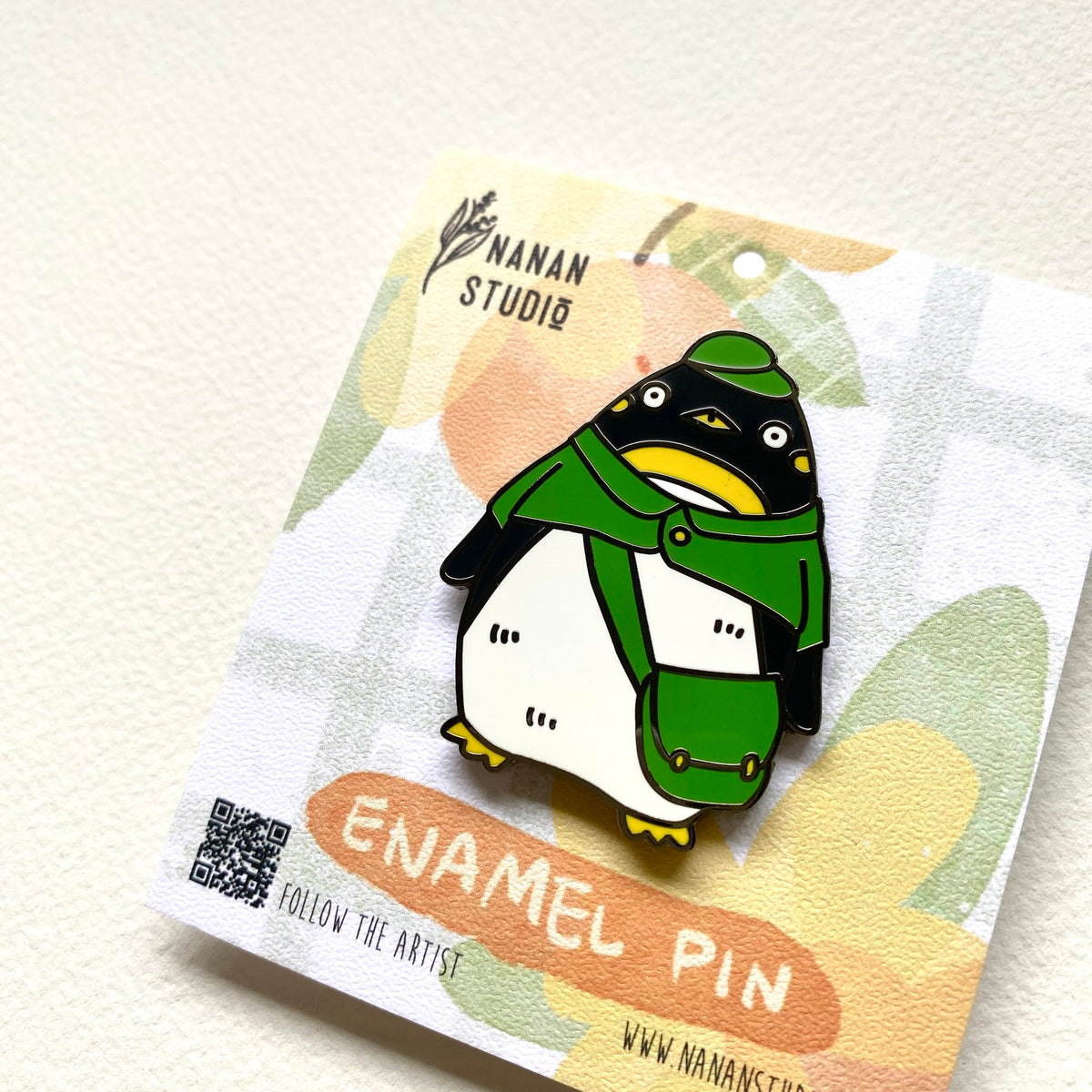 Close-up of the penguin postman enamel pin, showing the green cape, yellow beak and mail bag