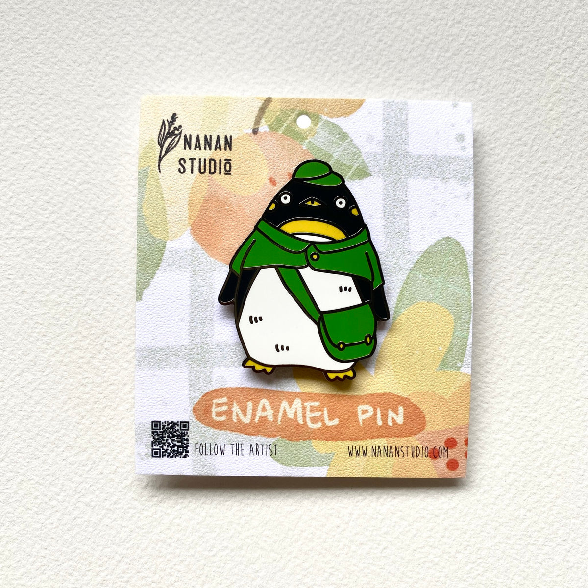 Penguin postman enamel pin in a green cap, green cape and mail bag, on an illustrated Nanan Studio backing card